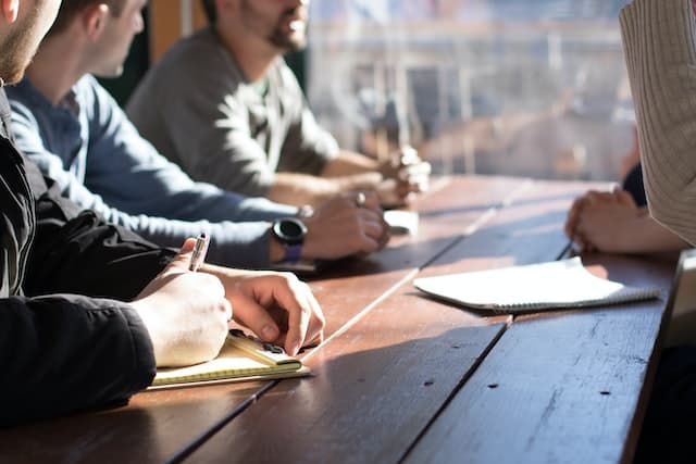 close up of a line of people resting their arms on a wooden table, holding notepads and pens