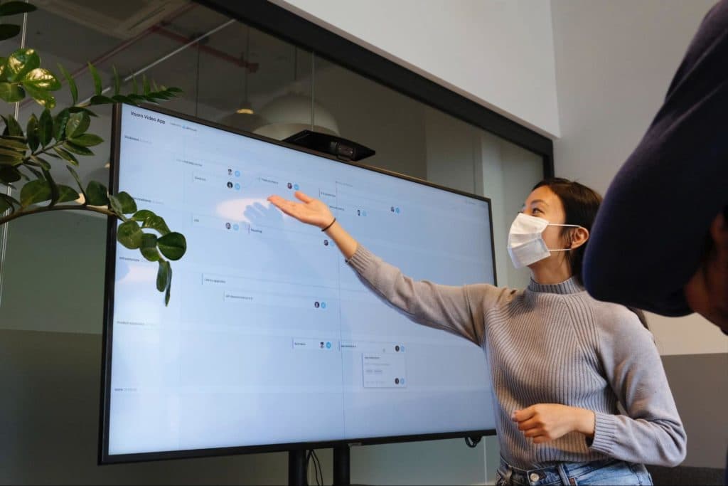 person wearing mask and gesturing to a roadmap laid out on a large screen behind them