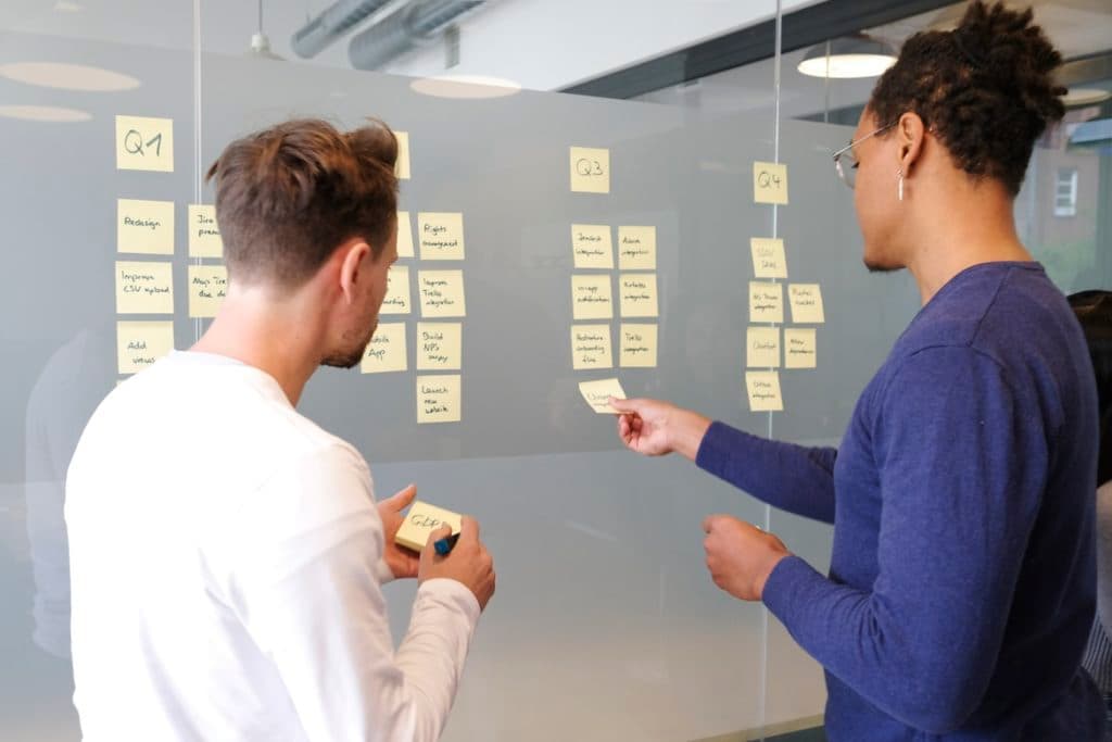 two people standing in front of glass office wall, with backs to the camera. they are looking at post-its stuck to the wall, broken down into the categories Q1, Q2, Q3, and Q4