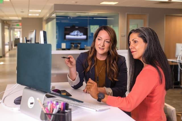 two people in an office setting sat in front of a laptop, both looking at the screen. one is pointing at the screen