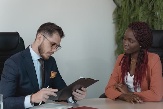 Two people sit at a table facing each other. One looks down at a clipboard